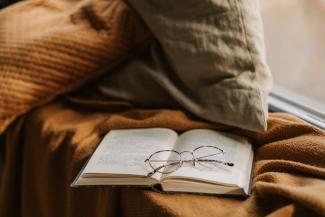 An open book on a windowsill. The book is resting on a knit blanket, and there is a pair of reading glasses on top of the book.